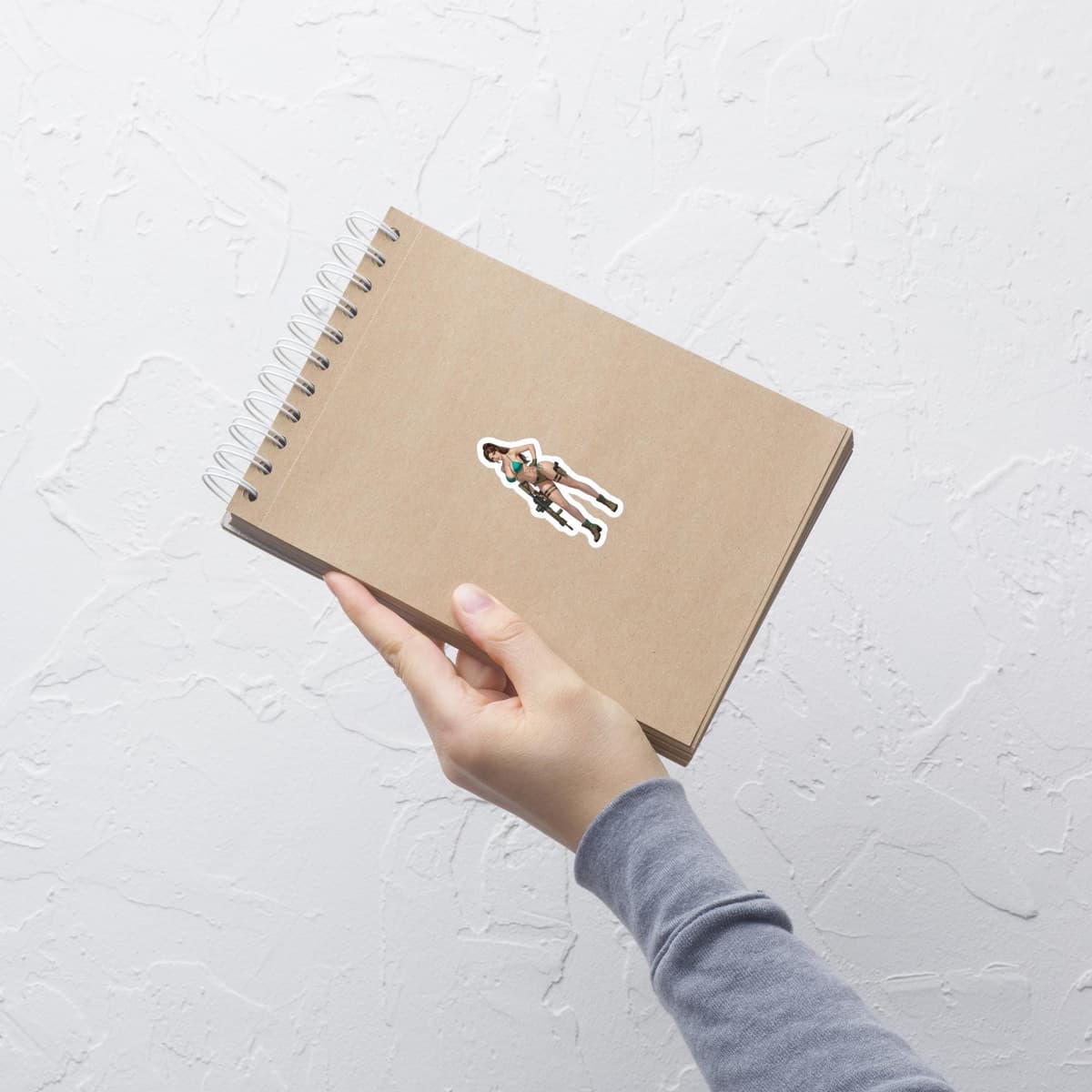May include: A sticker featuring a woman in a green bikini holding a rifle. The sticker is placed on a brown spiral-bound notebook. The notebook is held by a person wearing a gray long-sleeved shirt.