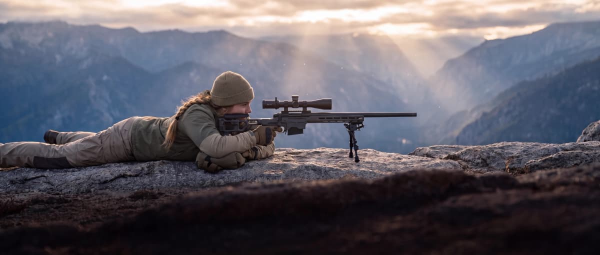 Prone shooter behind a bipod-supported bolt-action precision rifle on a granite ridgeline at golden hour
