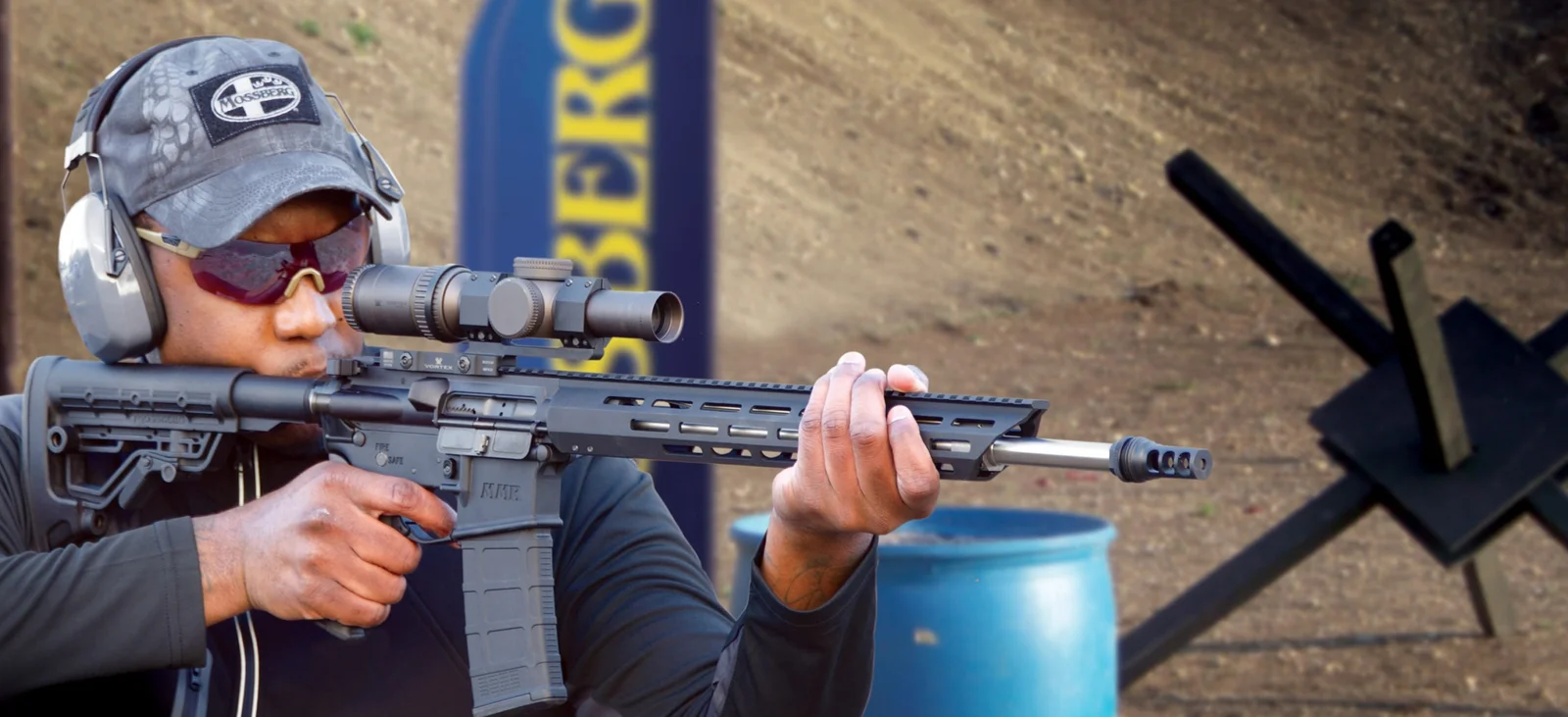 Shooter aiming a scoped AR-15-type rifle at an outdoor competition range