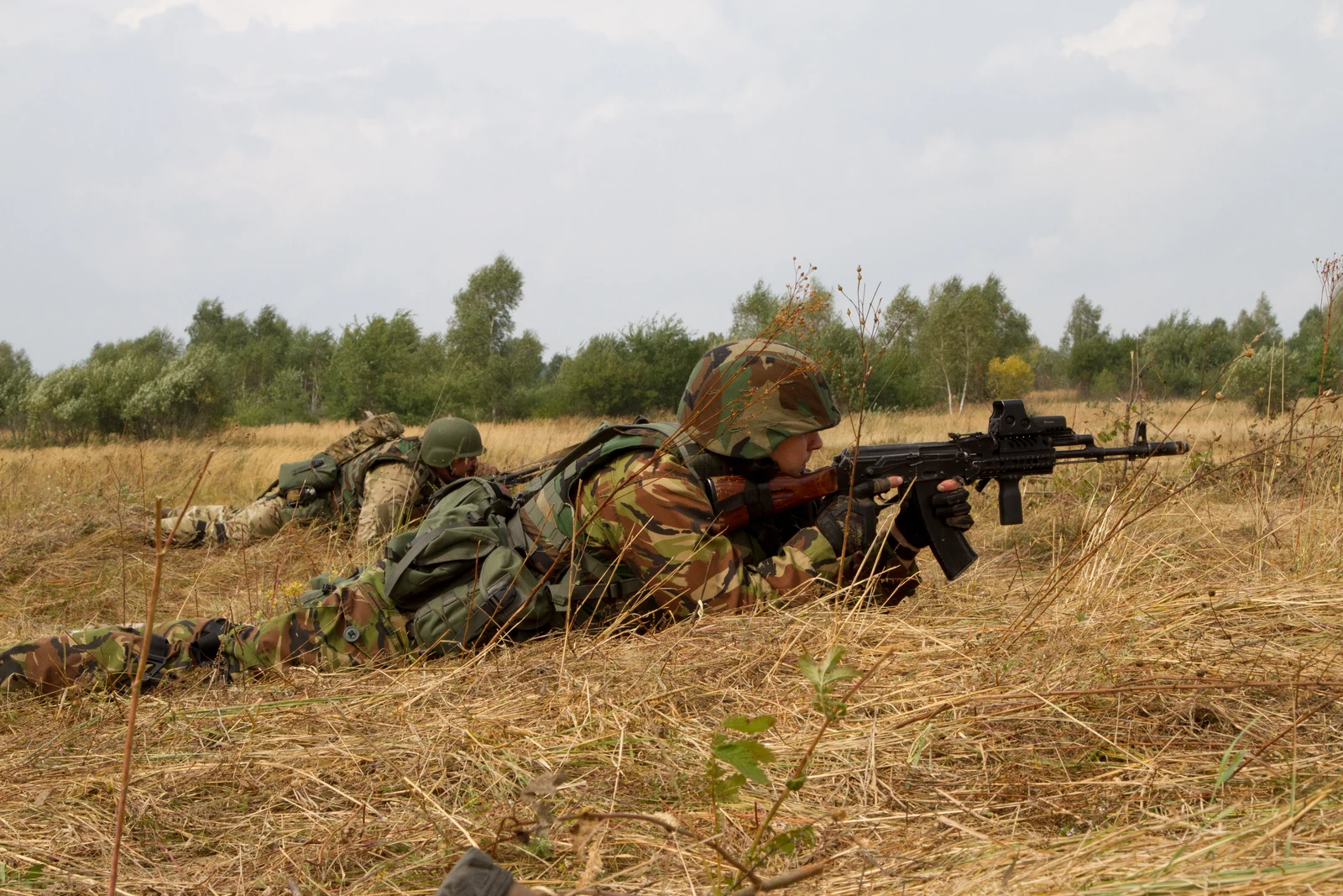 Ukrainian National Guard soldier prone in a field firing an AK-74 fitted with a railed aluminum handguard, a red dot sight, and a vertical foregrip, with an orange bakelite magazine