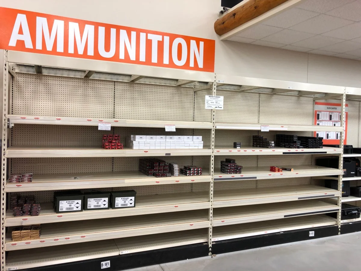 A nearly bare retail ammunition aisle under an orange AMMUNITION sign, with only scattered boxes left on otherwise empty shelves