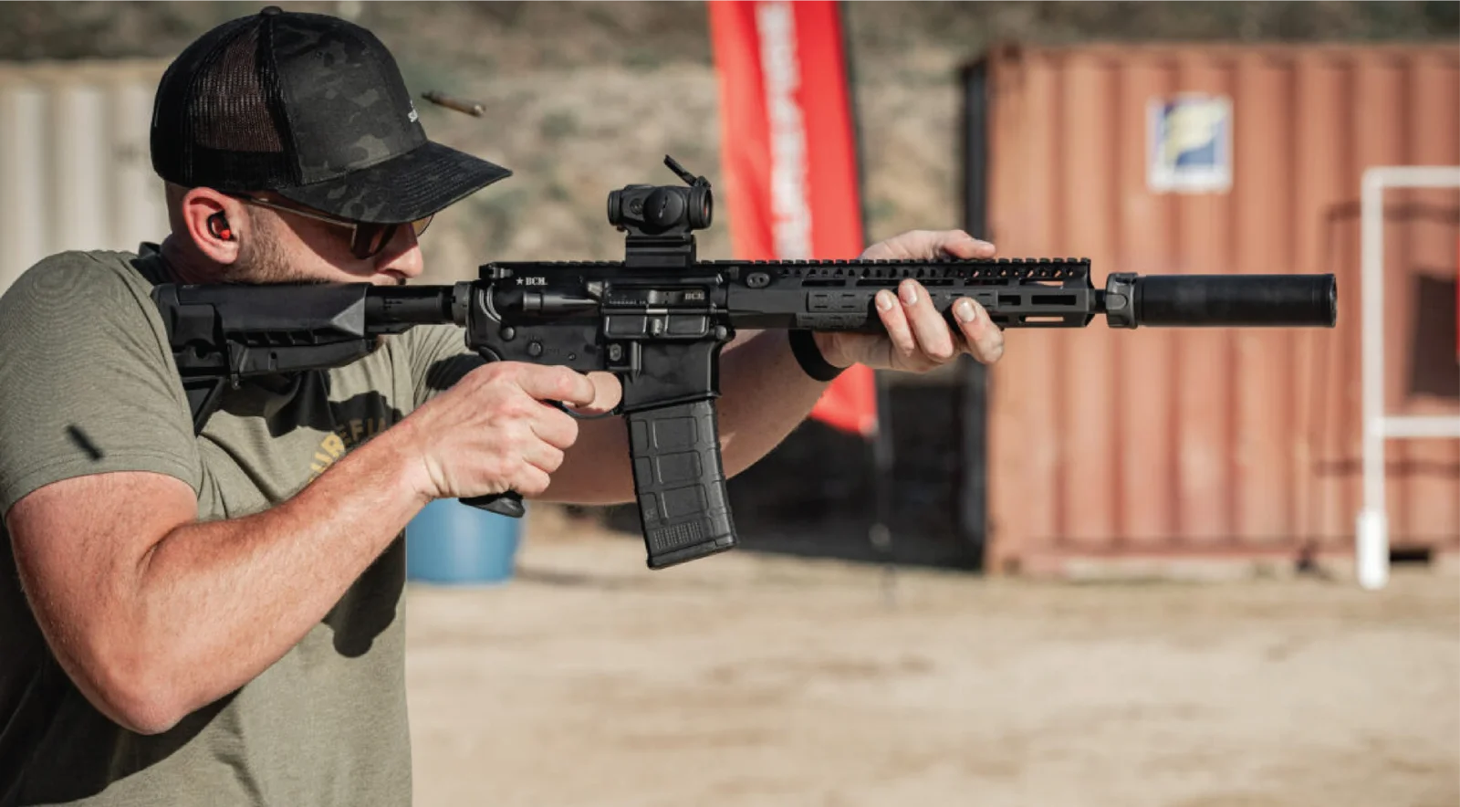Shooter firing a suppressed AR-15 carbine with a red dot and M-LOK handguard at an outdoor range