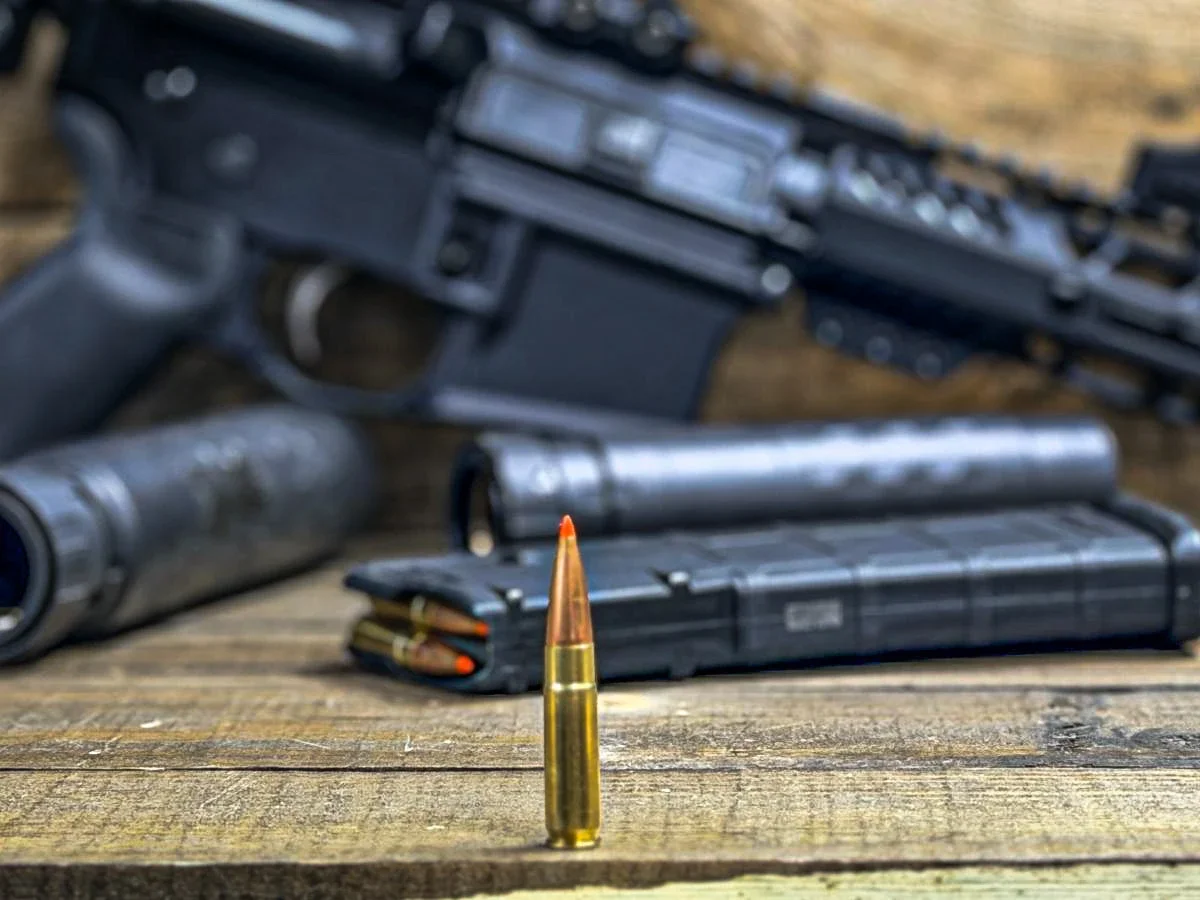 A .30-caliber rifle cartridge standing on a wood plank in front of a blurred AR-15, a suppressor, and loaded 30-round magazines