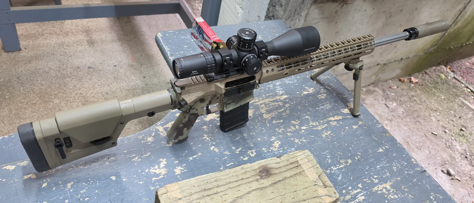 Arken EP-5 5-25x56 riflescope mounted on a tan and camouflage Aero Precision AR-10-pattern rifle with a suppressor and bipod on a range bench, a box of Hornady American Whitetail ammunition behind it