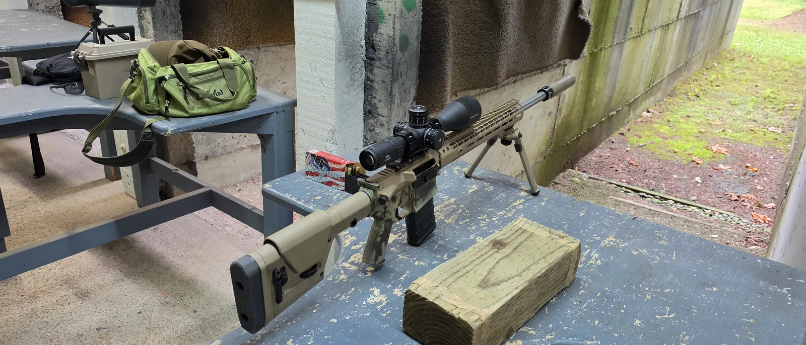 Arken EP-5 5-25x56 on a suppressed tan Aero Precision AR-10-pattern rifle resting on its bipod at a covered shooting bench, with a range bag and ammo can on the next bench