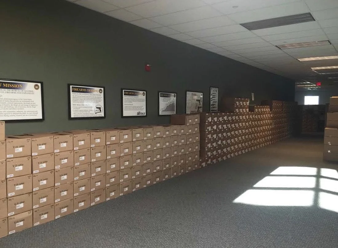 Corridor at the ATF National Tracing Center lined floor to shoulder height with stacked cardboard boxes of firearms records beneath framed ATF mission and firearms tracing posters