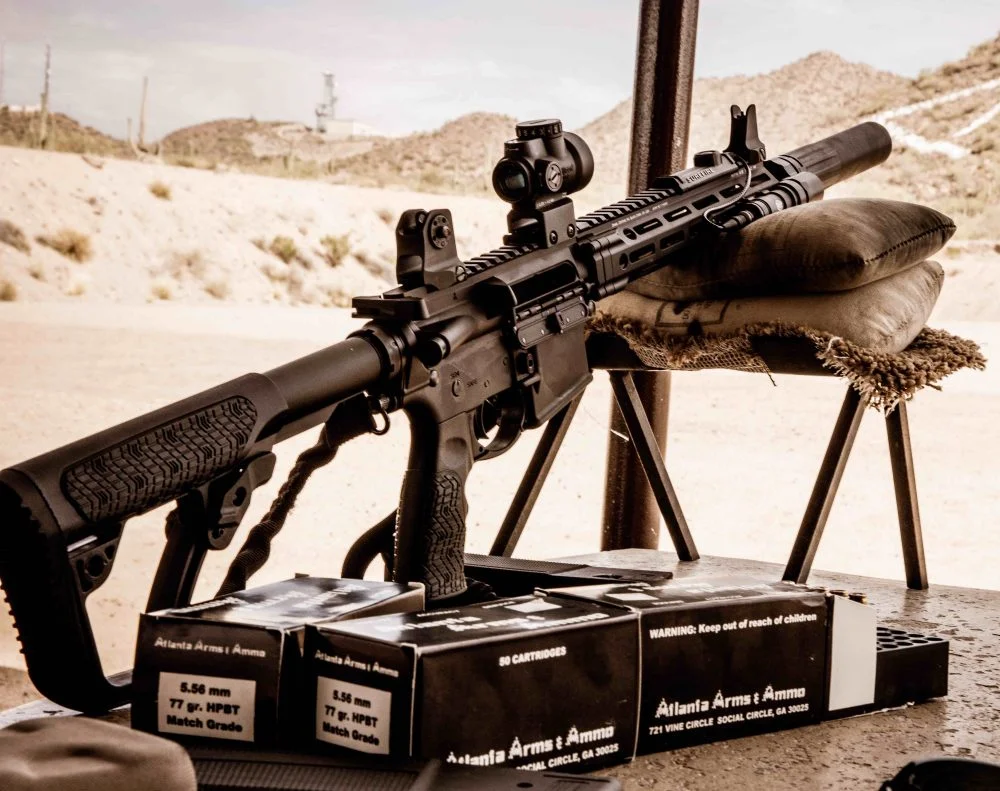 Suppressed AR-15 with a red dot and weapon light resting on sandbags at a desert range beside boxes of 5.56 ammunition