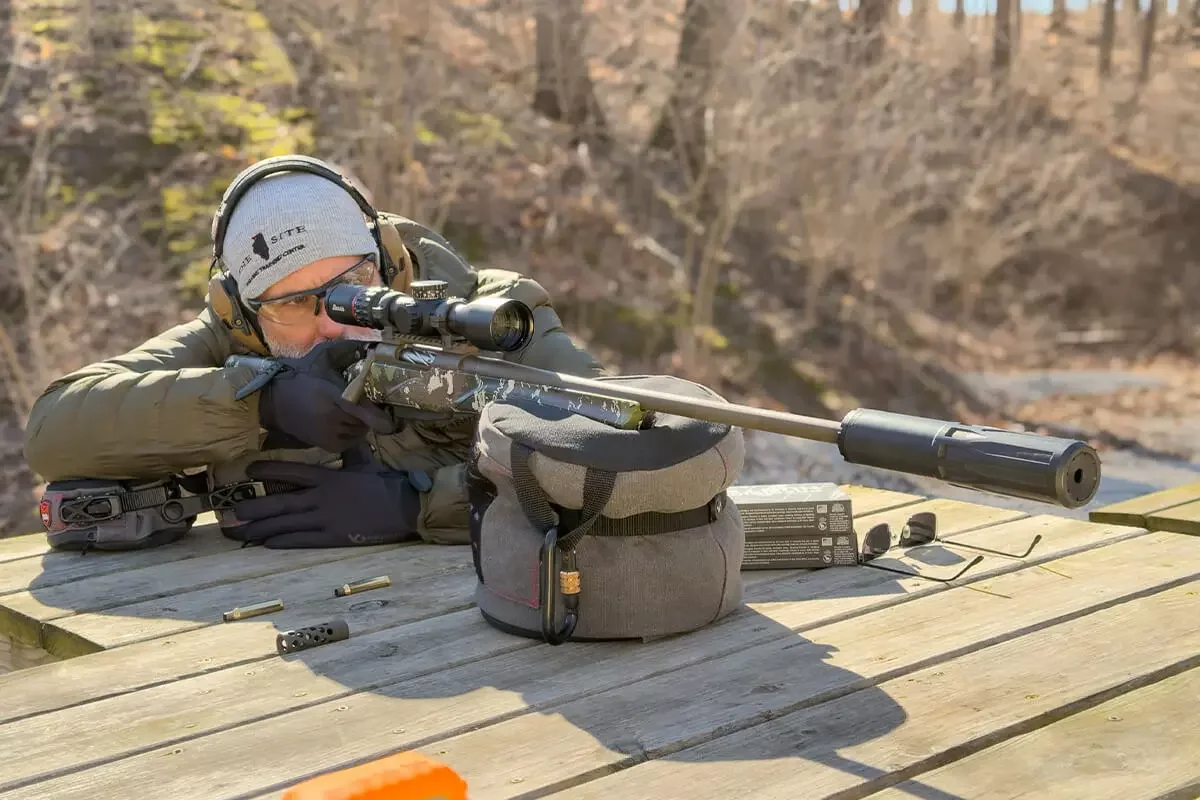 Shooter at bench aiming suppressed bolt-action rifle outdoors