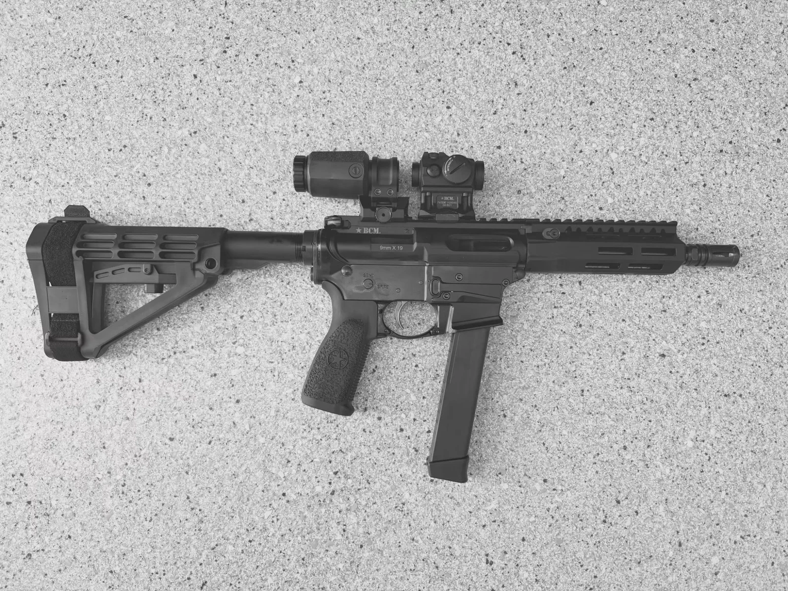 Kyle Defoor's BCM9 in profile on concrete, showing a braced 9mm AR with a red dot, a magnifier, an M-LOK handguard, and a polymer Glock-pattern magazine