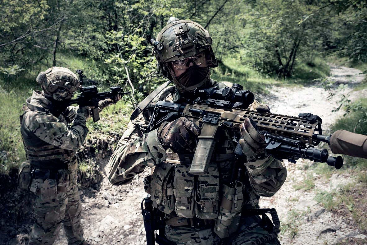 Two soldiers in multicam on a wooded trail, the lead soldier holding a tan Beretta NARP rifle with a magnified optic and a suppressor