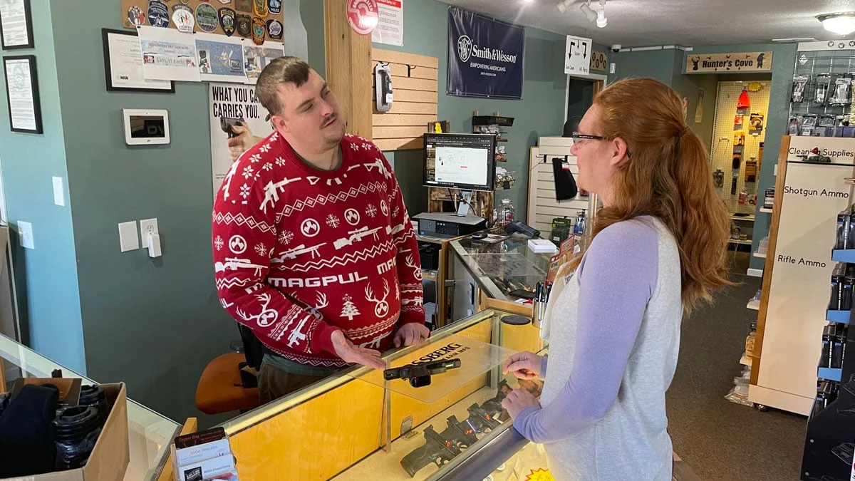 A gun shop employee talking with a customer across a glass display counter stocked with handguns