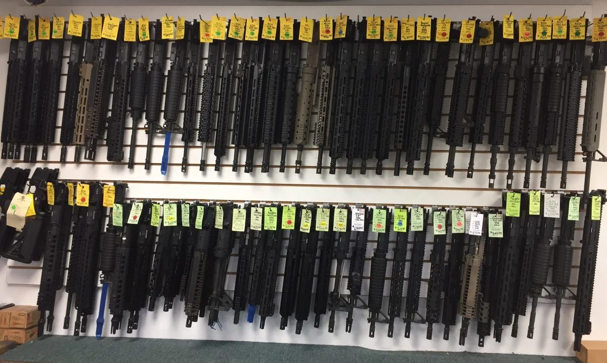 Two racked rows of complete AR-15 upper receivers with barrels, each tagged with a handwritten price label on a gun shop wall