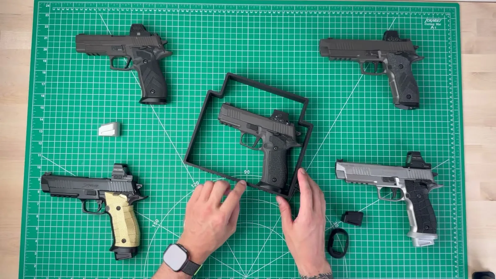A SIG P226 sitting inside a rectangular gun-test box gauge on a cutting mat, with four other P226 variants and a raw aluminum magwell laid out around it