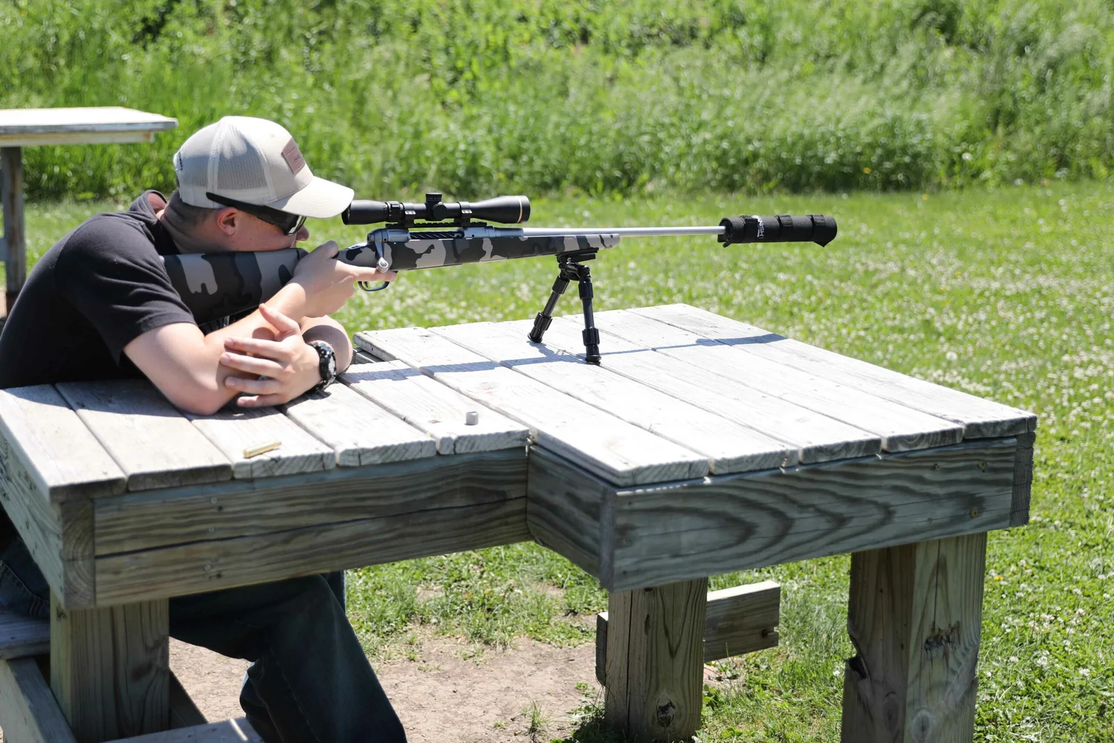 Shooter firing a suppressed camouflage bolt-action rifle from a wooden bench at an outdoor range