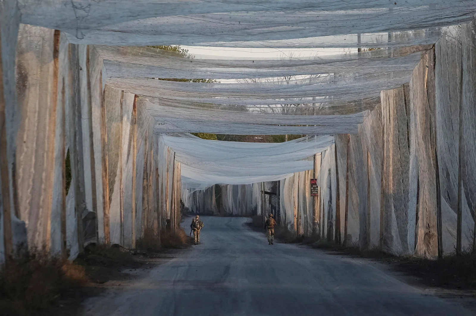 Two soldiers walking down a rural road covered end to end by an anti-drone netting tunnel strung from tall poles on both sides