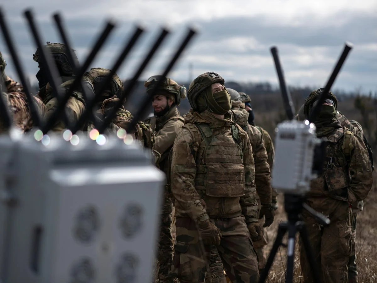 Soldiers in a field standing beside tripod-mounted electronic warfare jammers with clusters of vertical antennas, one soldier scanning the sky overhead