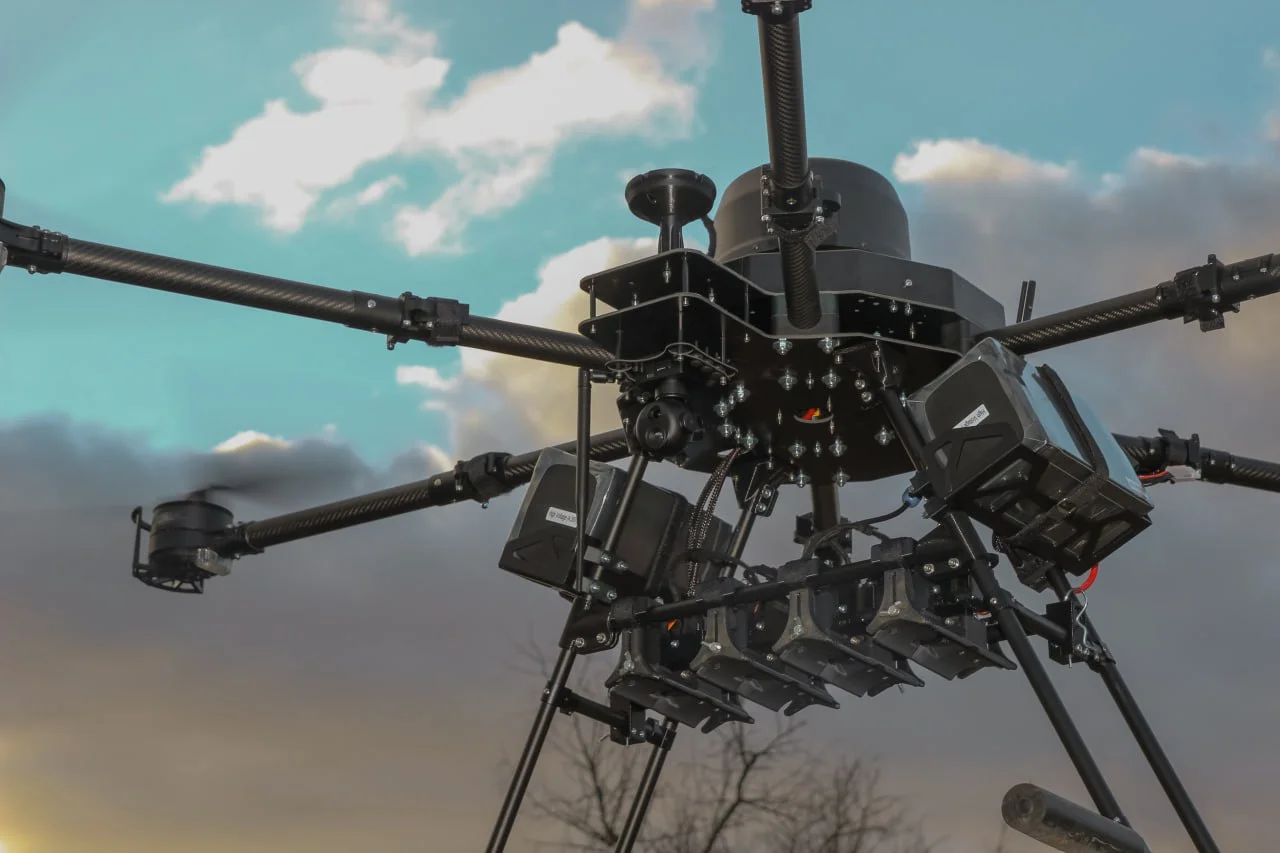 Underside of a heavy multirotor bomber drone in flight, showing a carbon frame, a gimballed camera, battery packs, and a rack of finned drop munitions slung beneath the airframe