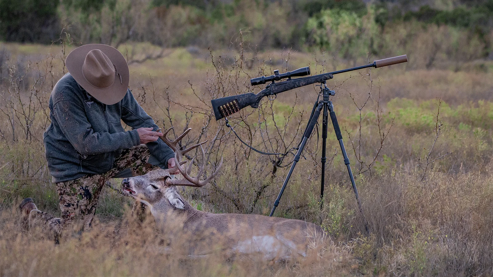 Hunter with bolt-action rifle on a tripod next to a harvested whitetail buck in a grassy field