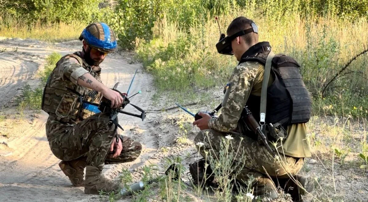 Two Ukrainian soldiers preparing an FPV strike drone, one holding the quadcopter and the other wearing FPV goggles with a hobby-style radio transmitter