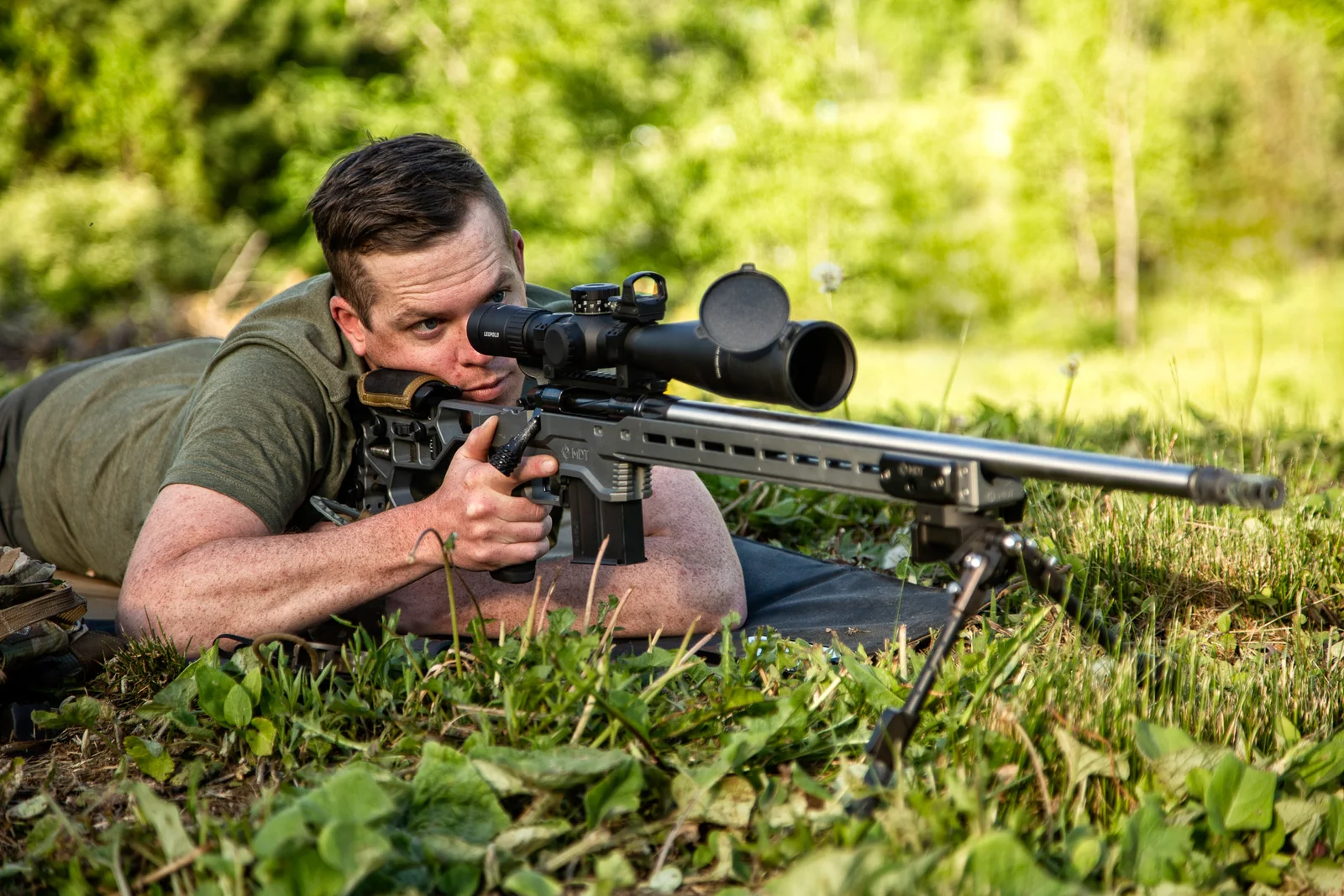 Shooter prone behind a chassis-stocked bolt-action rifle with a Leupold scope and bipod in a grass field