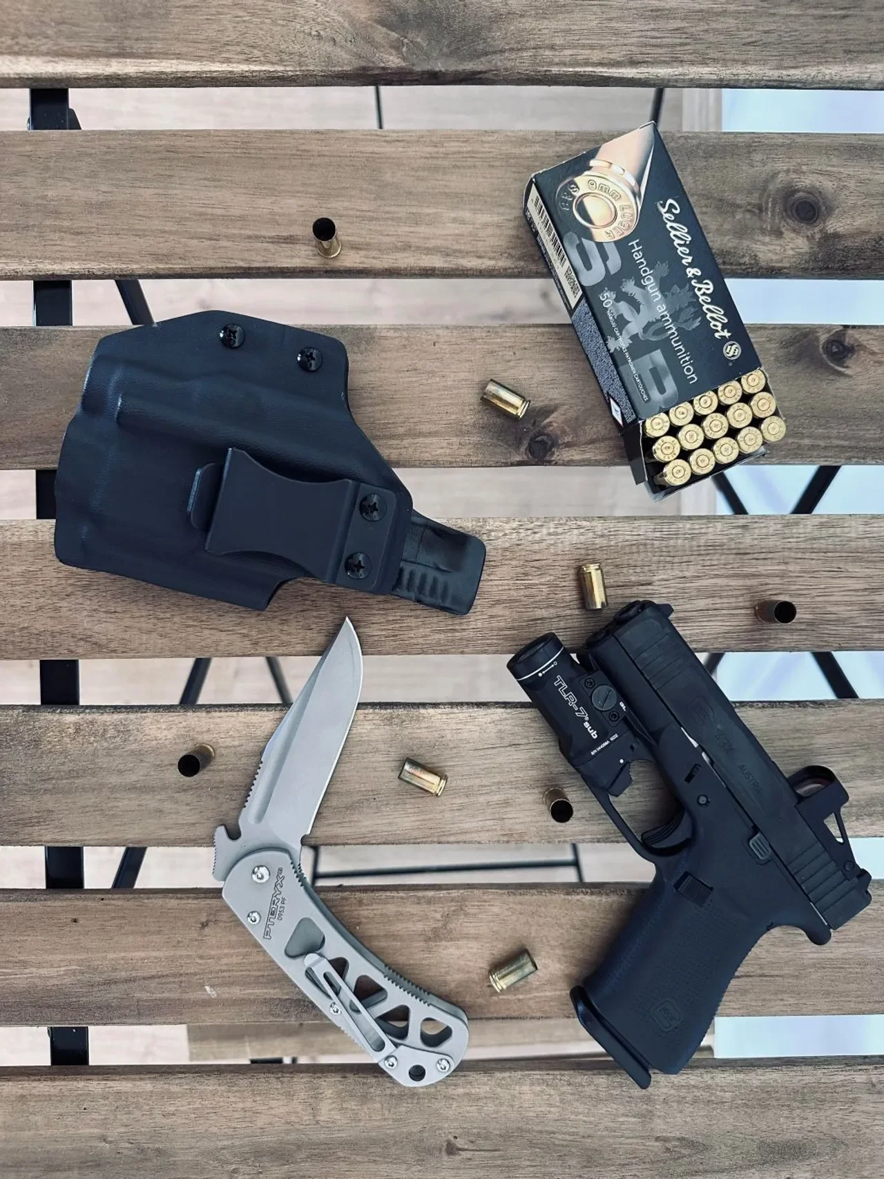 Everyday carry flatlay with a kydex holster, Glock with weapon light, folding knife, and 9mm ammunition