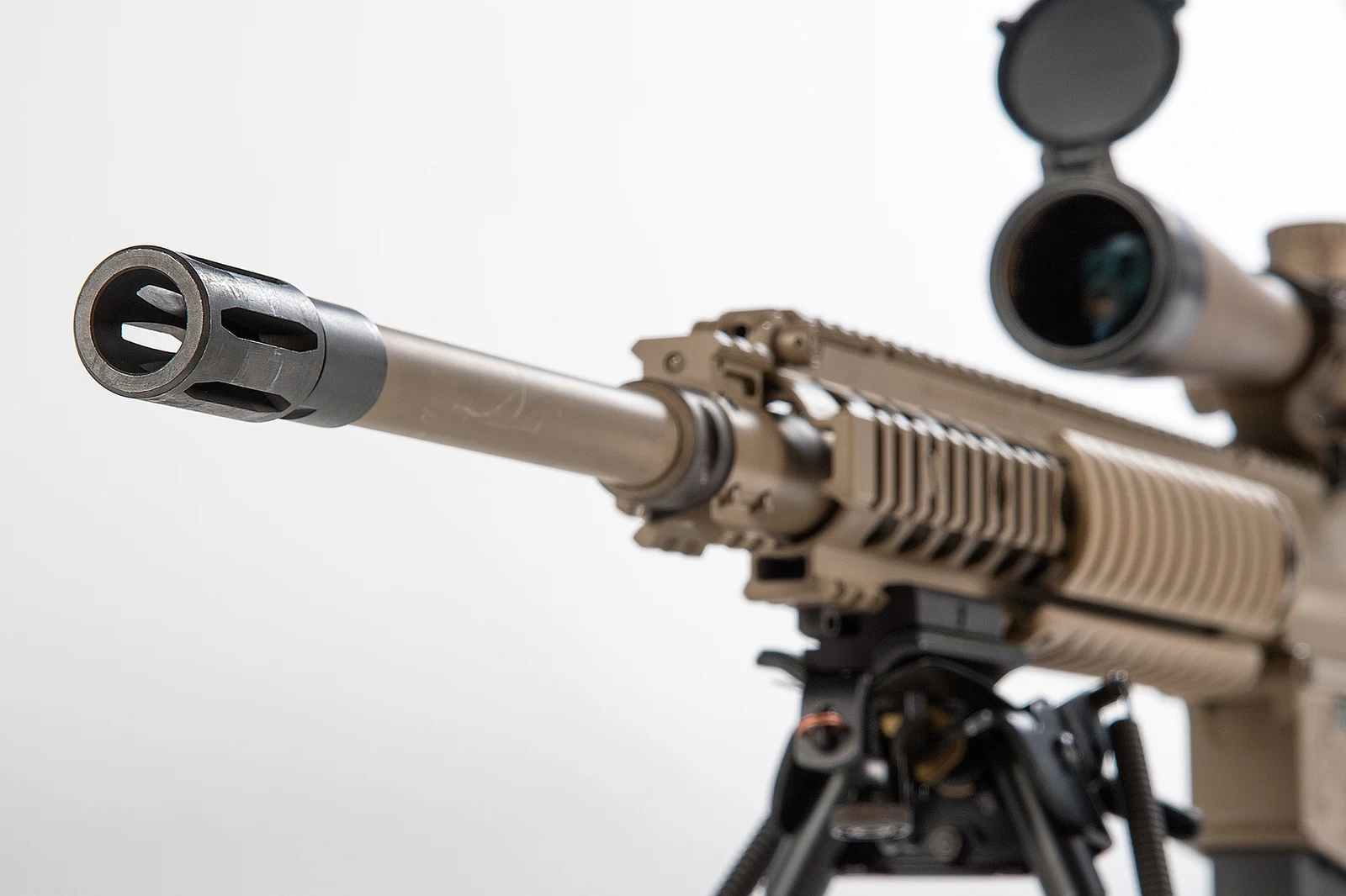 Close view of an M110 Semi-Automatic Sniper System muzzle, flash suppressor, and free-float rail section with a scope and bipod behind it