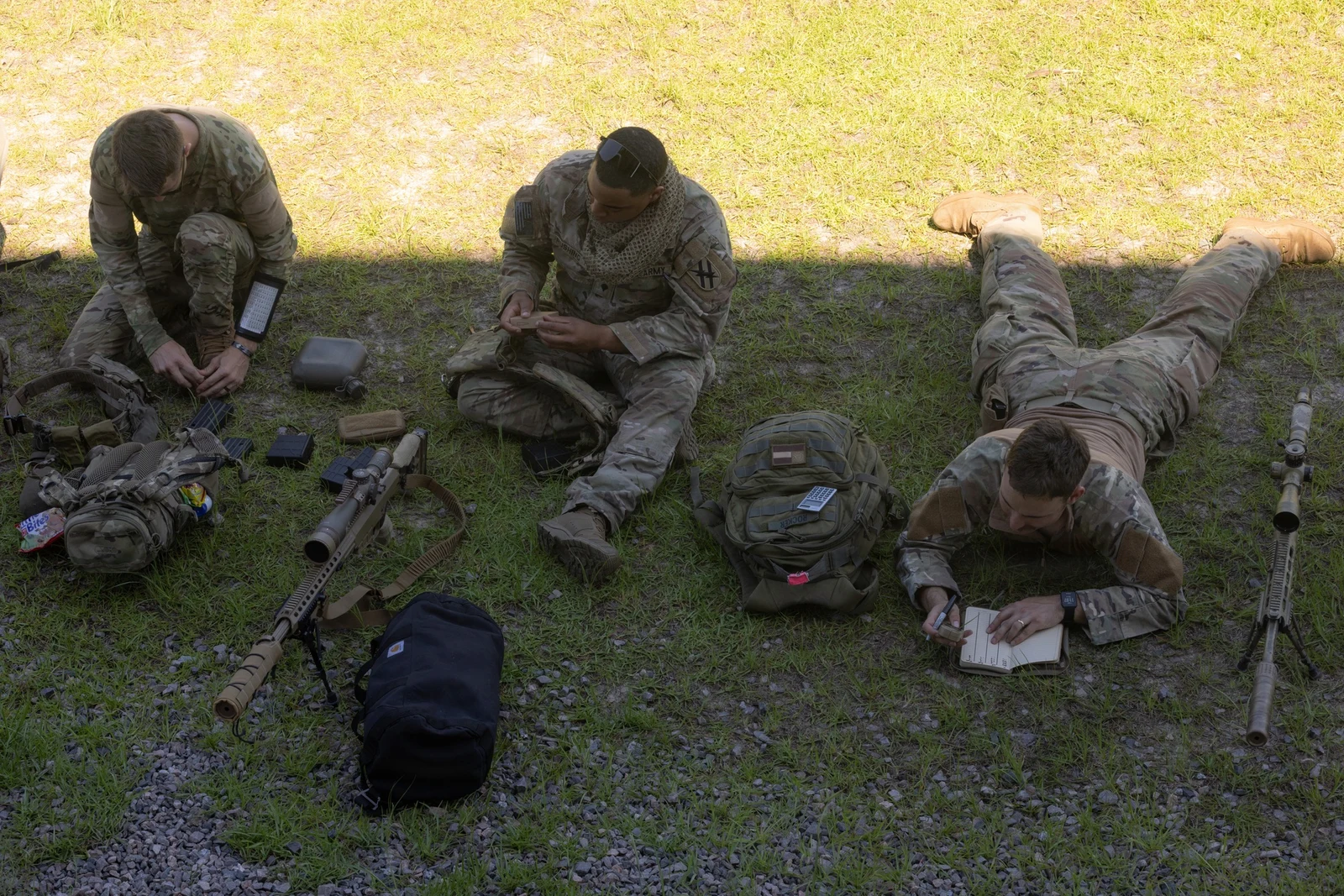 Two suppressed M110 rifles laid out on the grass beside soldiers recording data in dope books during annual training