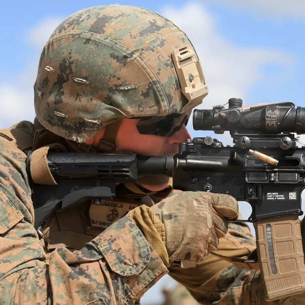 A US Marine in MARPAT uniform and helmet aiming an M4 carbine fitted with a Trijicon ACOG
