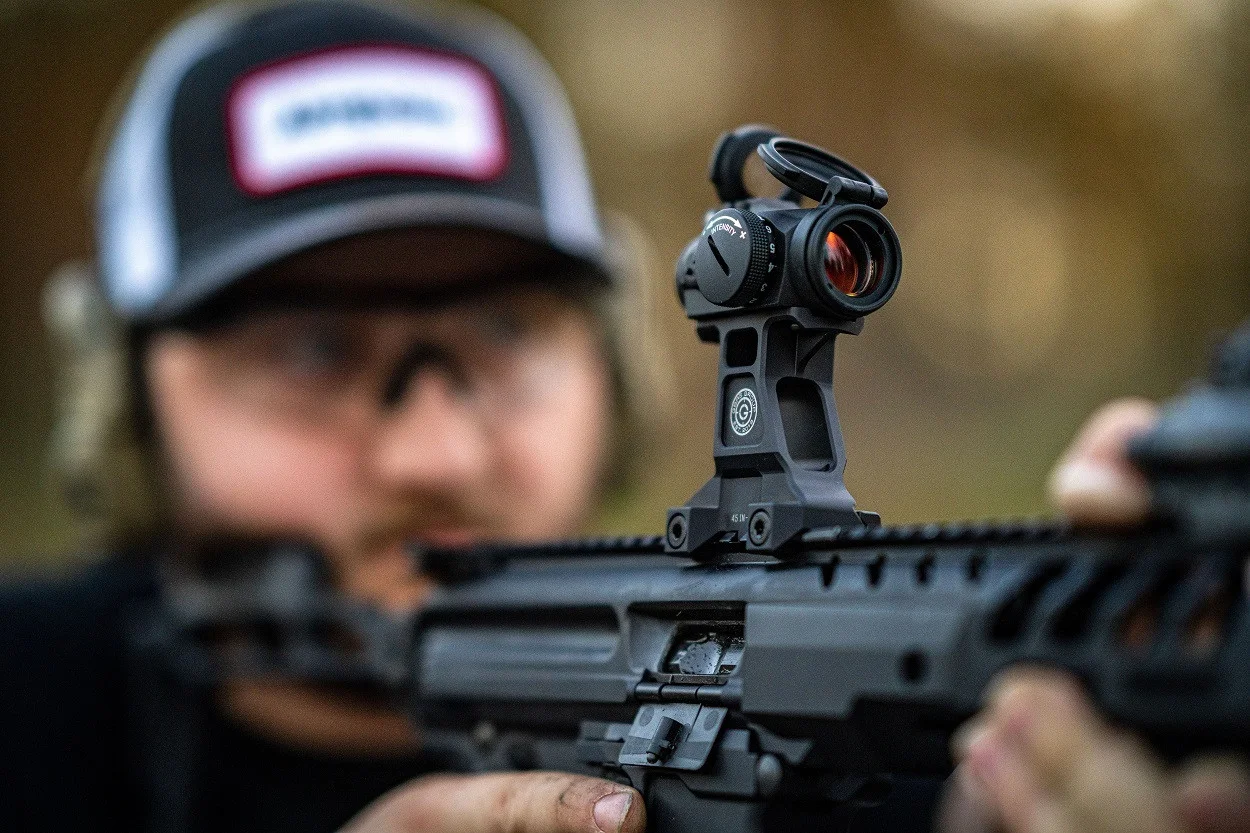 An Aimpoint micro red dot sight on a riser mount atop an AR-15 upper receiver, with a shooter behind it