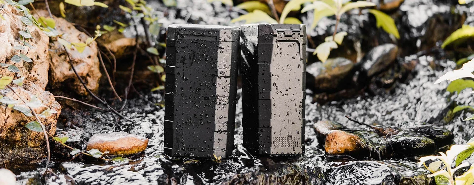 Two Magpul DAKA Can 2.0 polymer containers standing in shallow running water on wet rocks, beaded with water droplets