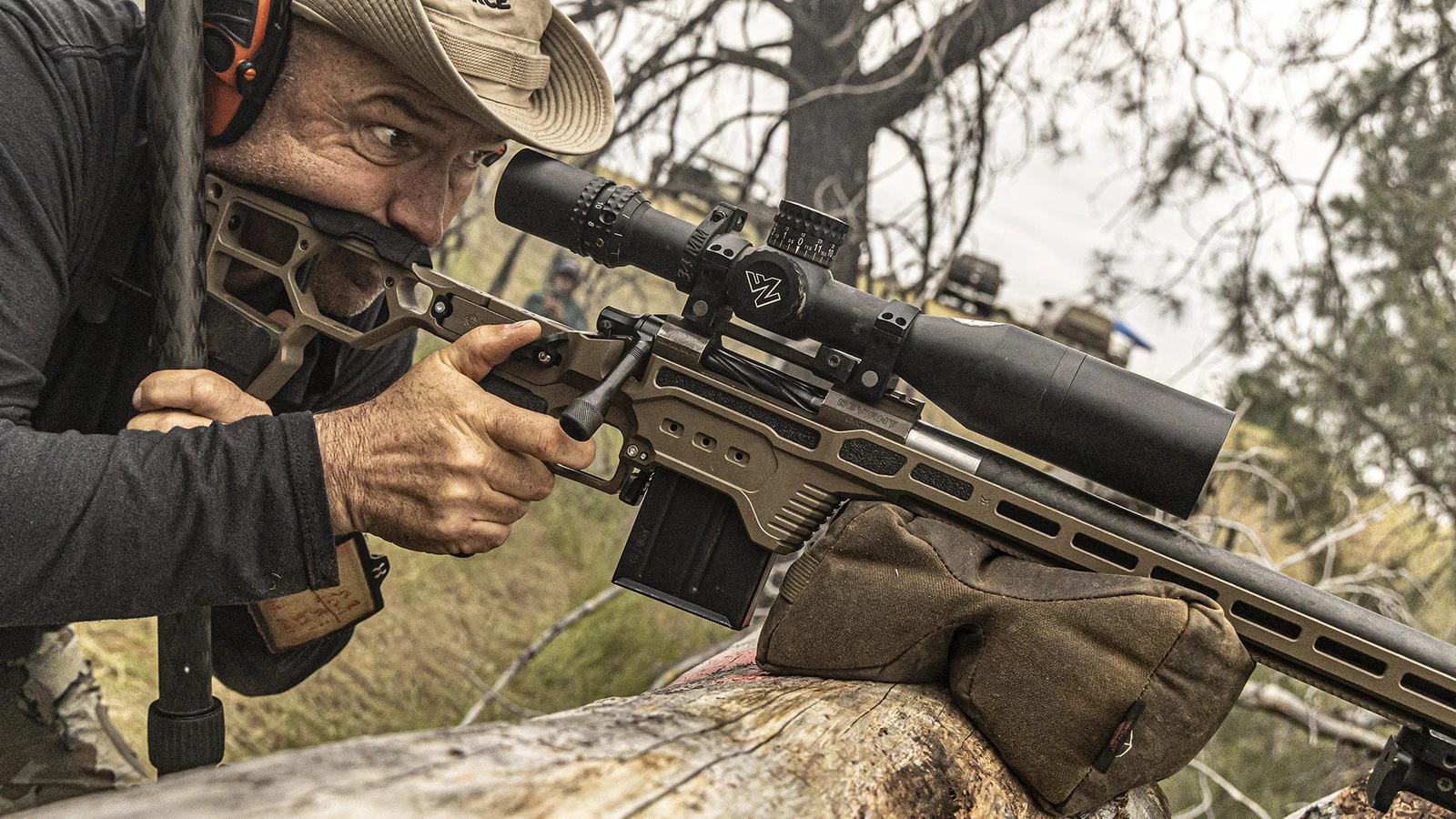 Shooter firing an FDE MDT DRT-chassis rifle from a rear bag rested on a log