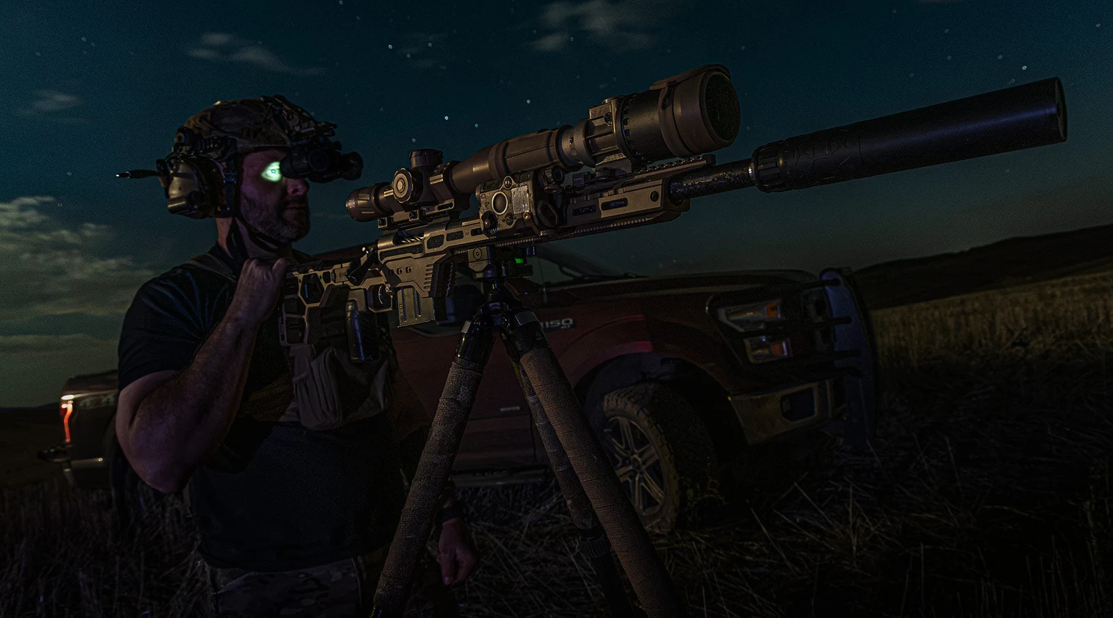 Hunter wearing a night vision device standing beside a DRT-chassis rifle on a tripod at night, with a clip-on device mounted ahead of the scope