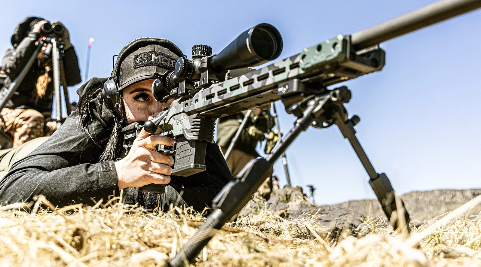 Shooter prone behind a camo-finished MDT chassis rifle on a bipod, with a scope, AICS magazine, and MDT cap, at an outdoor range