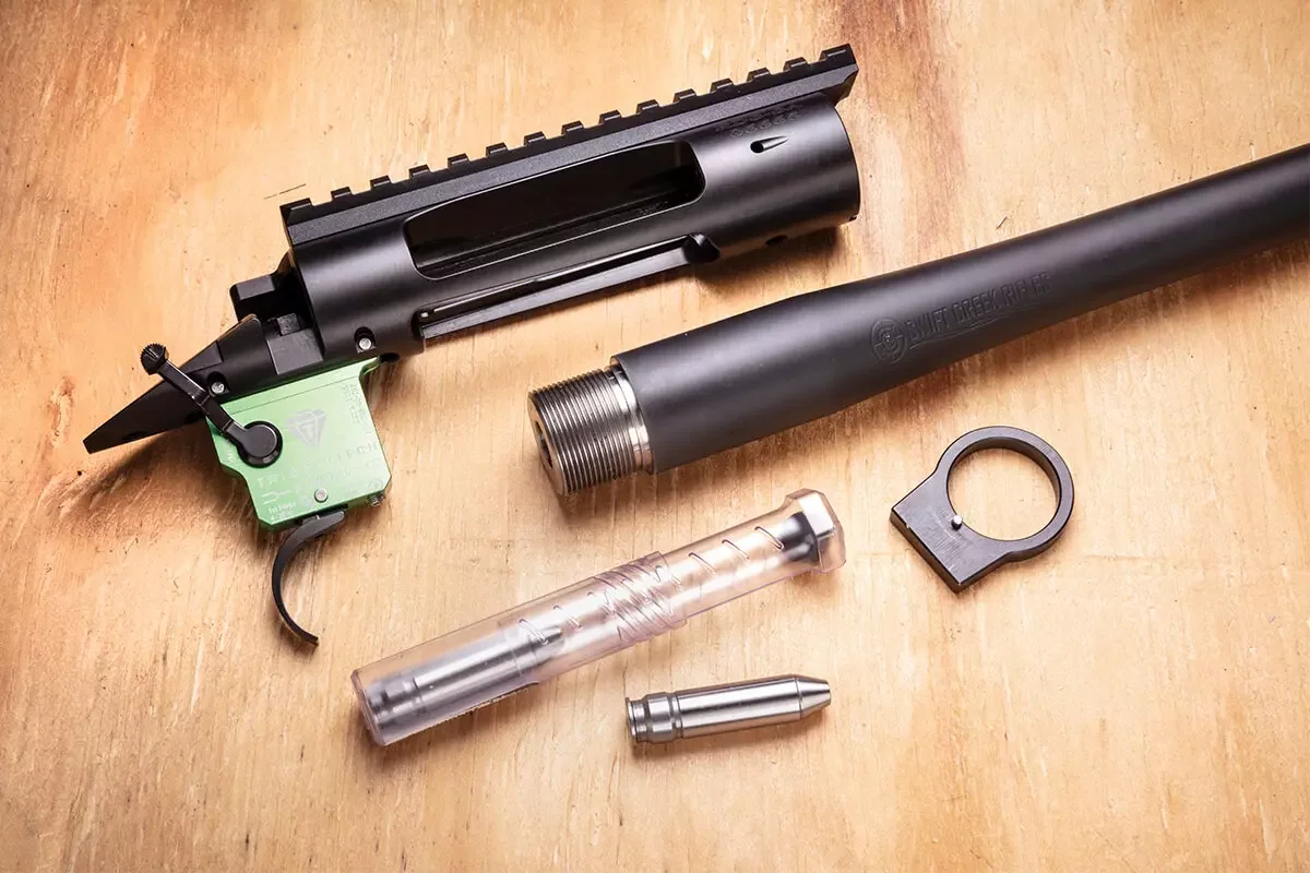 Disassembled custom bolt action rifle parts on a wooden bench: black receiver with rail, threaded barrel blank, green TriggerTech trigger, recoil lug, and bolt