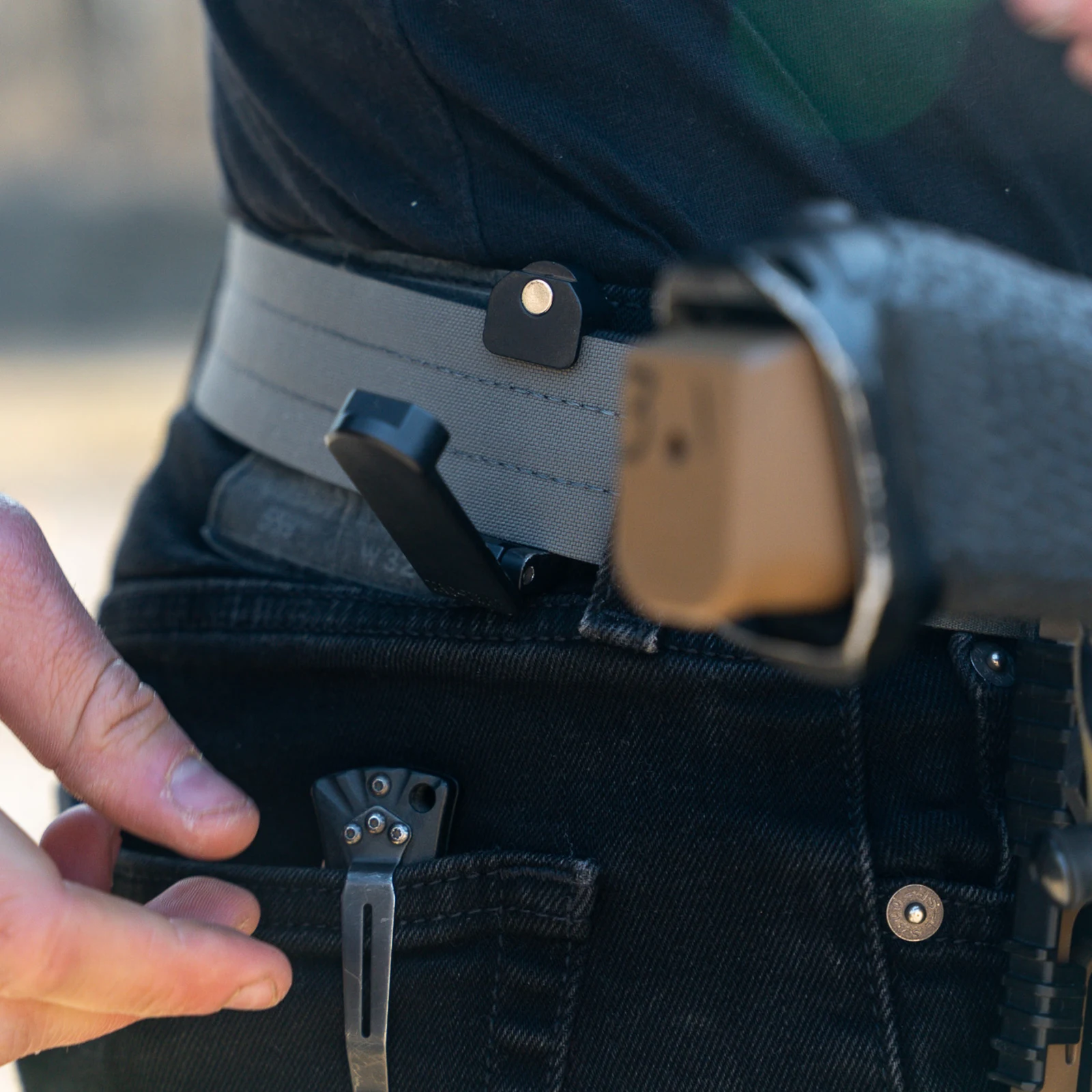 A NeoMag Tac-Trap magnetic retainer mounted on a light gray belt at the waistline, with a hand reaching toward a pocket-clipped knife and a tan magazine carried alongside