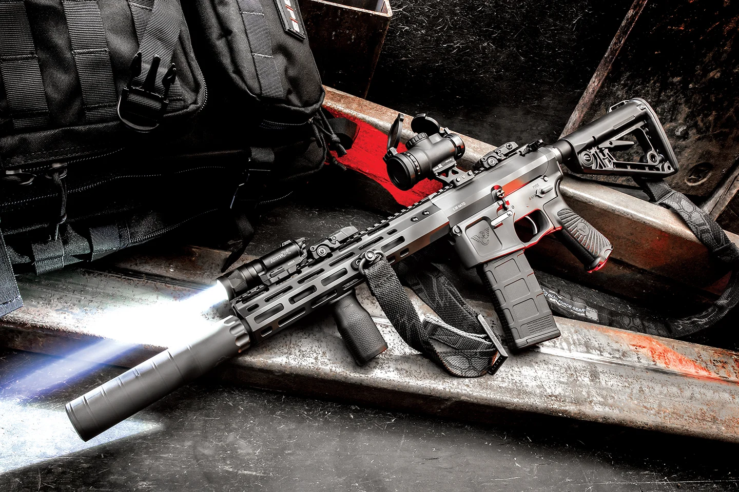 A suppressed short-barreled AR-15 with a stock, red dot, and weapon light staged on steel