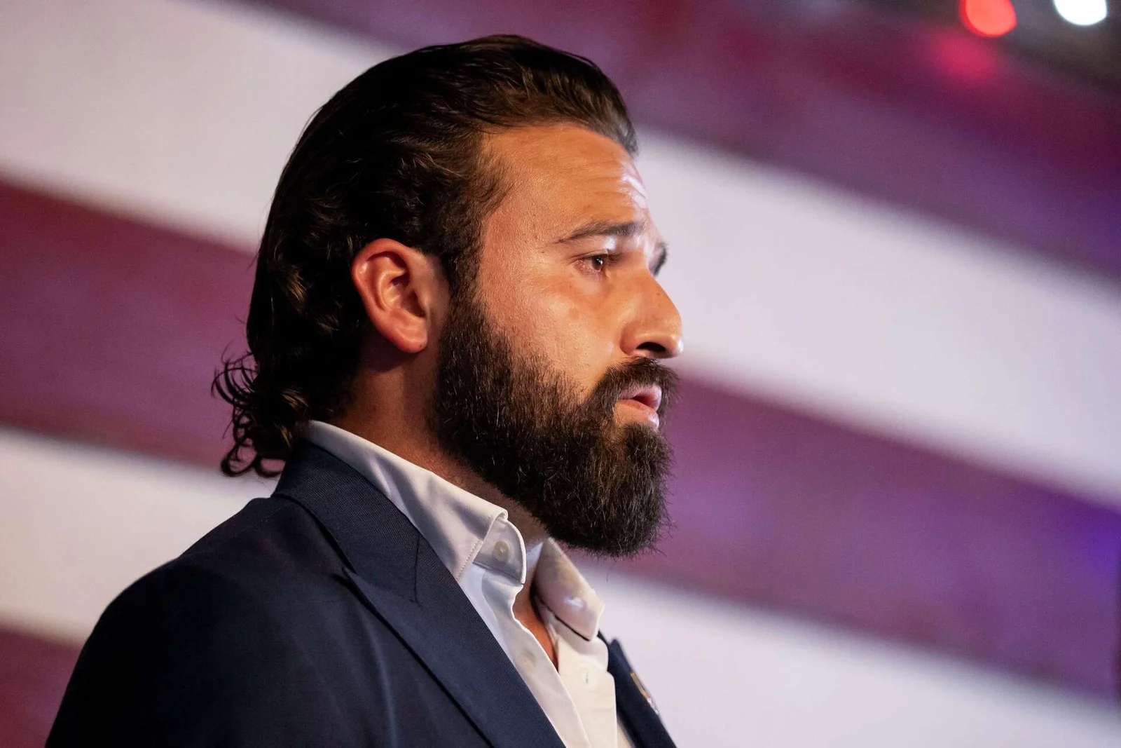 Brandon Herrera in a navy suit speaking in front of an American flag
