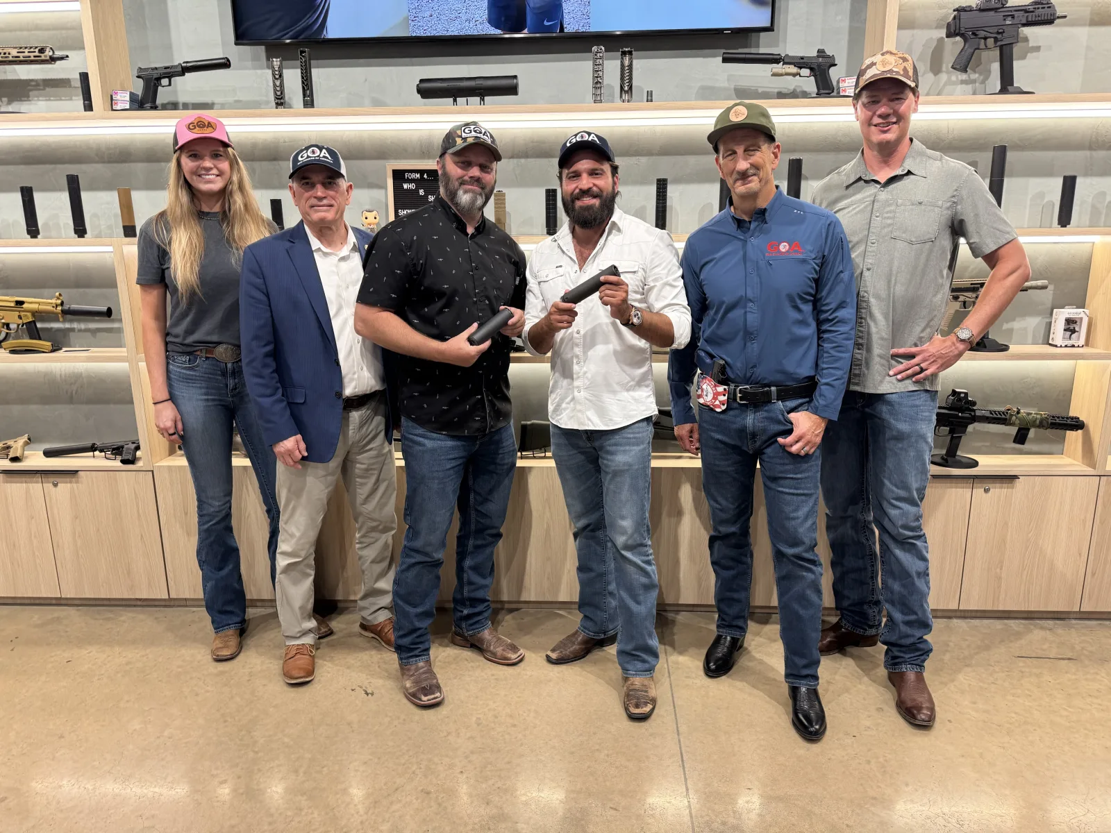 Six people in GOA hats standing in front of a lit wall of suppressors and suppressed firearms at the Silencer Shop storefront, two of them holding black suppressors
