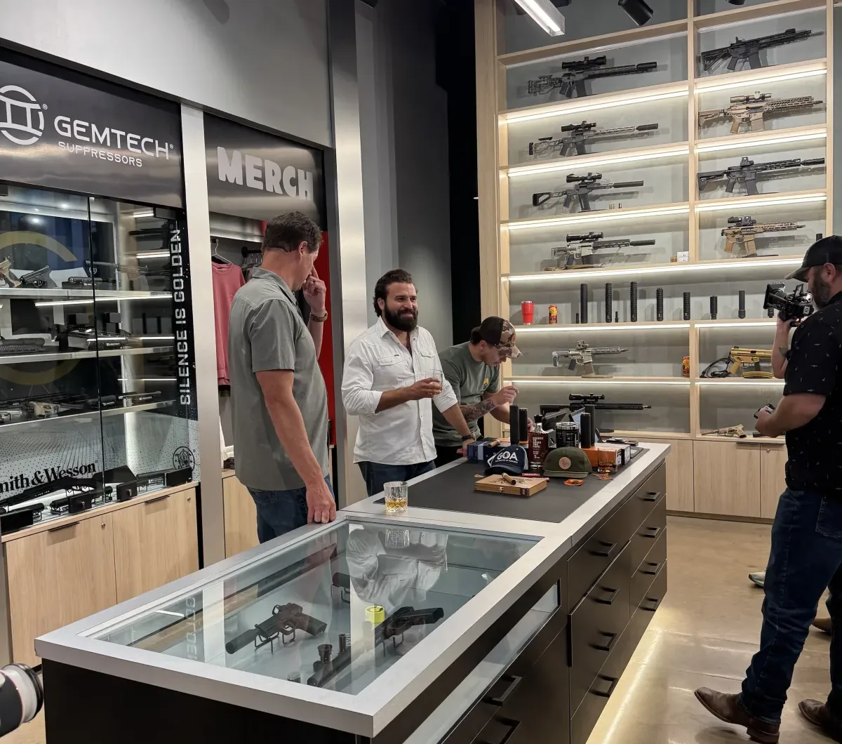 Interior of the Silencer Shop storefront at night, with a lit wall of suppressed rifles behind the sales counter and a GOA hat, whiskey, and cigars staged on the glass case
