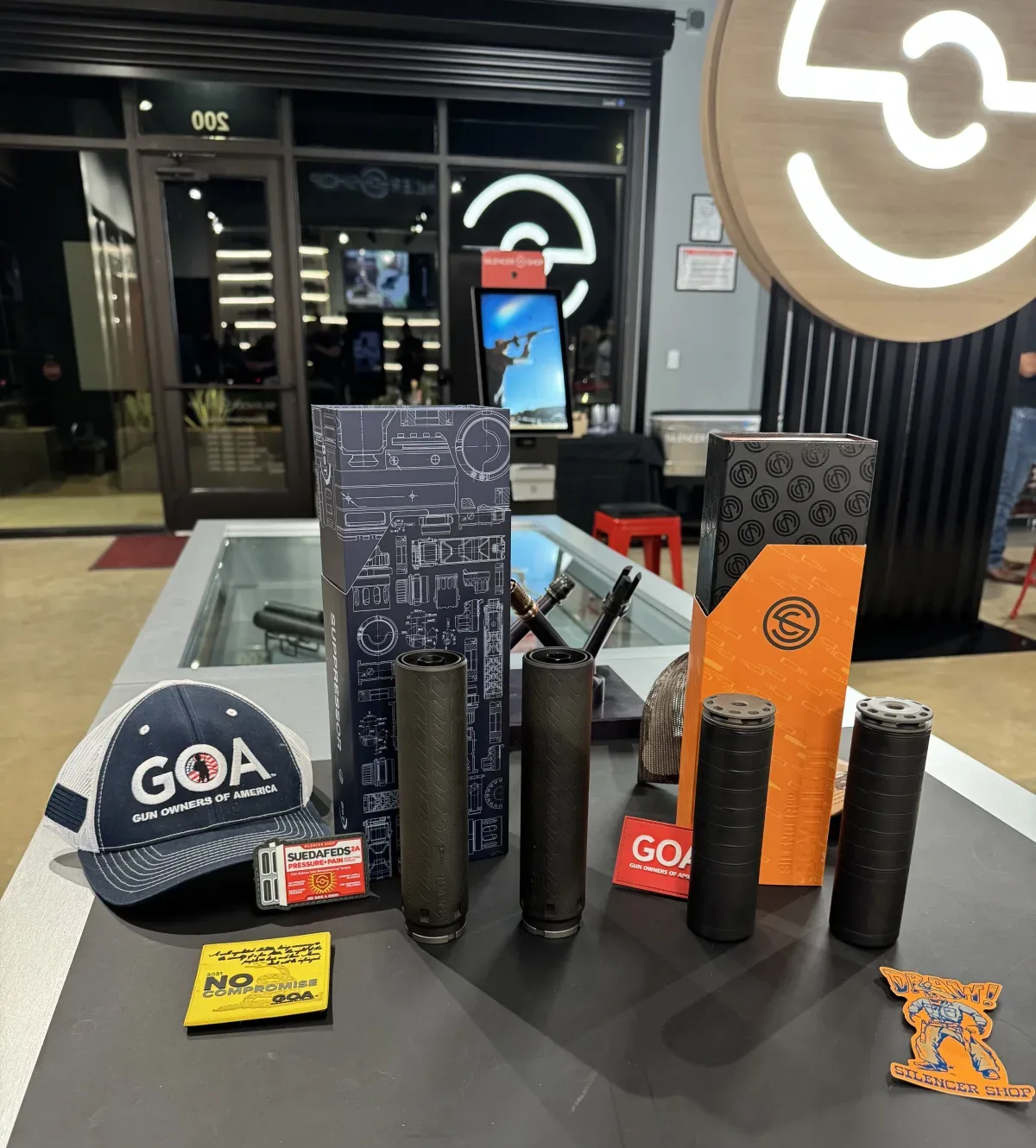 Suppressors and their manufacturer boxes staged on a counter outside the Silencer Shop storefront, alongside a GOA hat and a No Compromise handbook