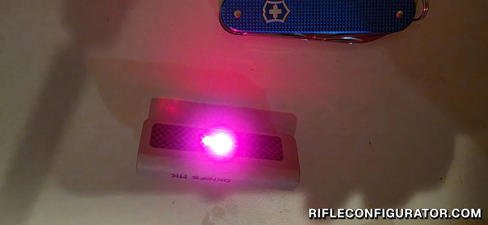 OKNIFE MK lying on a table with its red light on, next to a Victorinox pocket knife