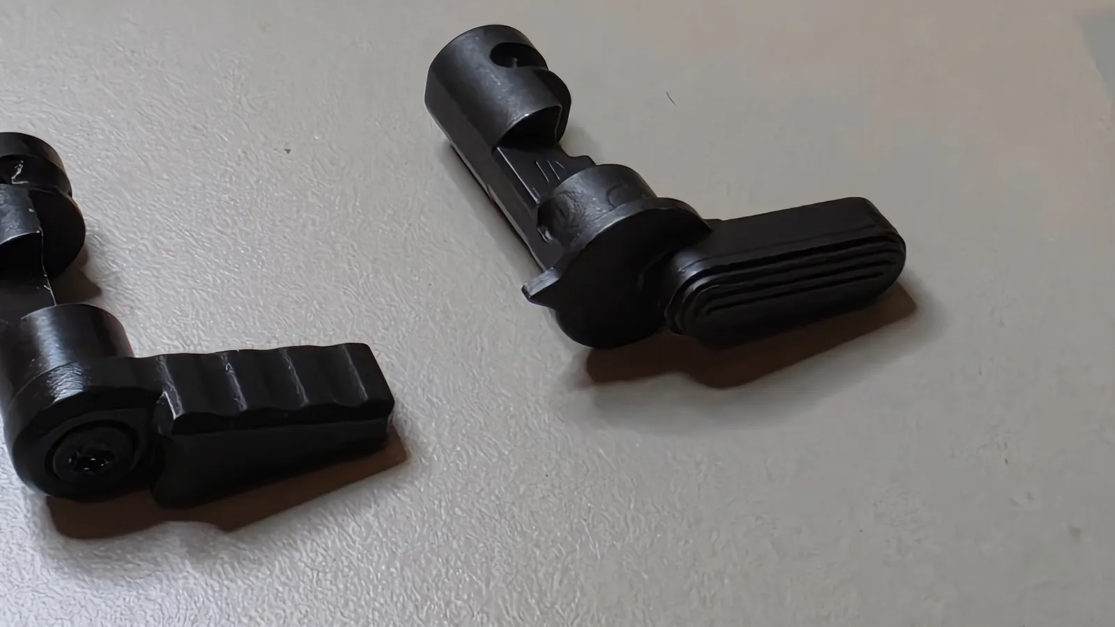 Two Triggered Company safety selectors on a bench: the launch-era two-piece selector with a serrated screw-on lever on the left and the OOPS one-piece selector with a lined mil-spec-style lever on the right