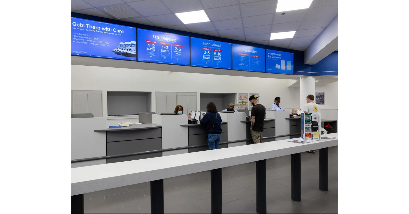 Customers at the retail counter of a U.S. Post Office lobby with USPS shipping menu screens overhead