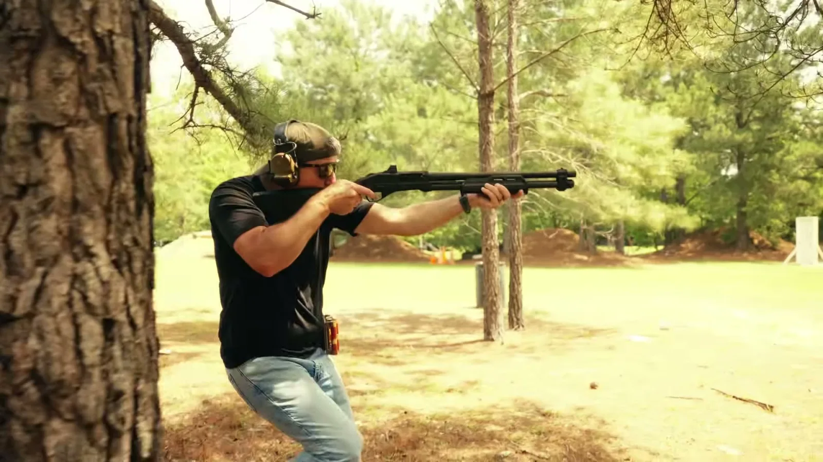 A shooter fires a black PSA 570 pump shotgun with a mounted red dot at an outdoor wooded range
