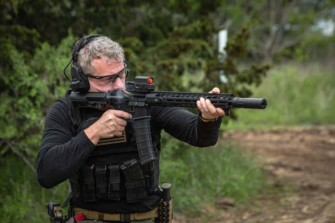 Shooter firing a suppressed AR-pattern rifle with a red dot optic at an outdoor range
