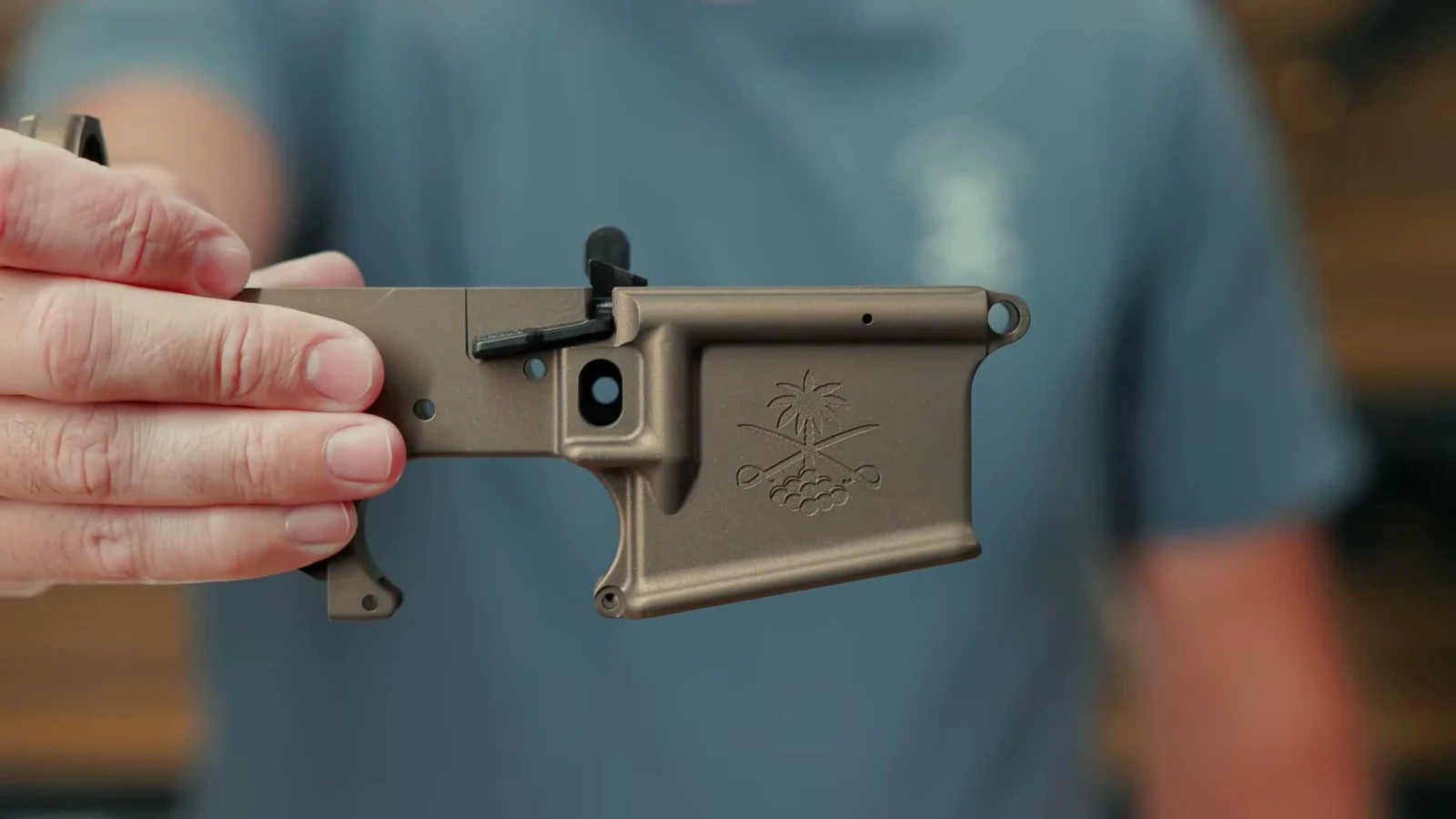 Burnt Bronze PSA Sabre Ambi Mil-Spec lower receiver held in hand, showing the black right-side ambidextrous bolt catch paddle above the magwell