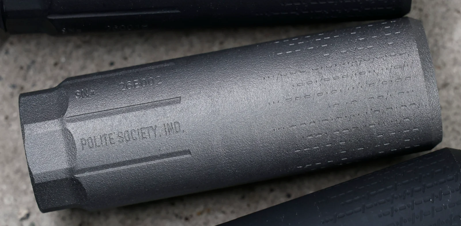 Polite Society Fortitude 5.56 Ti suppressor on concrete, showing POLITE SOCIETY, IND. marking, serial, wrench flats, and dashed texture toward the muzzle