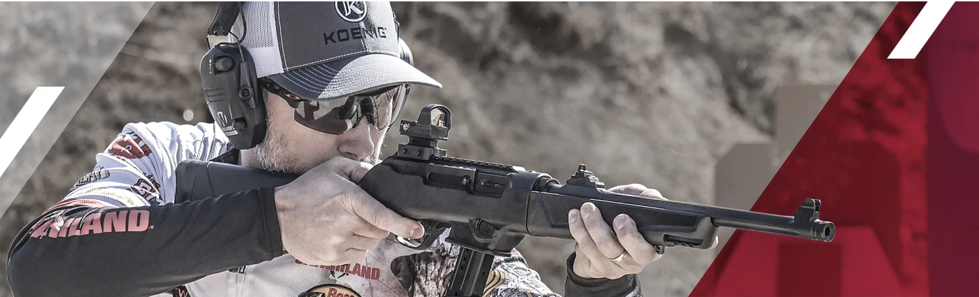 Shooter in a Koenig cap and electronic ear protection firing a black Ruger PC Carbine with a red dot sight on an outdoor range