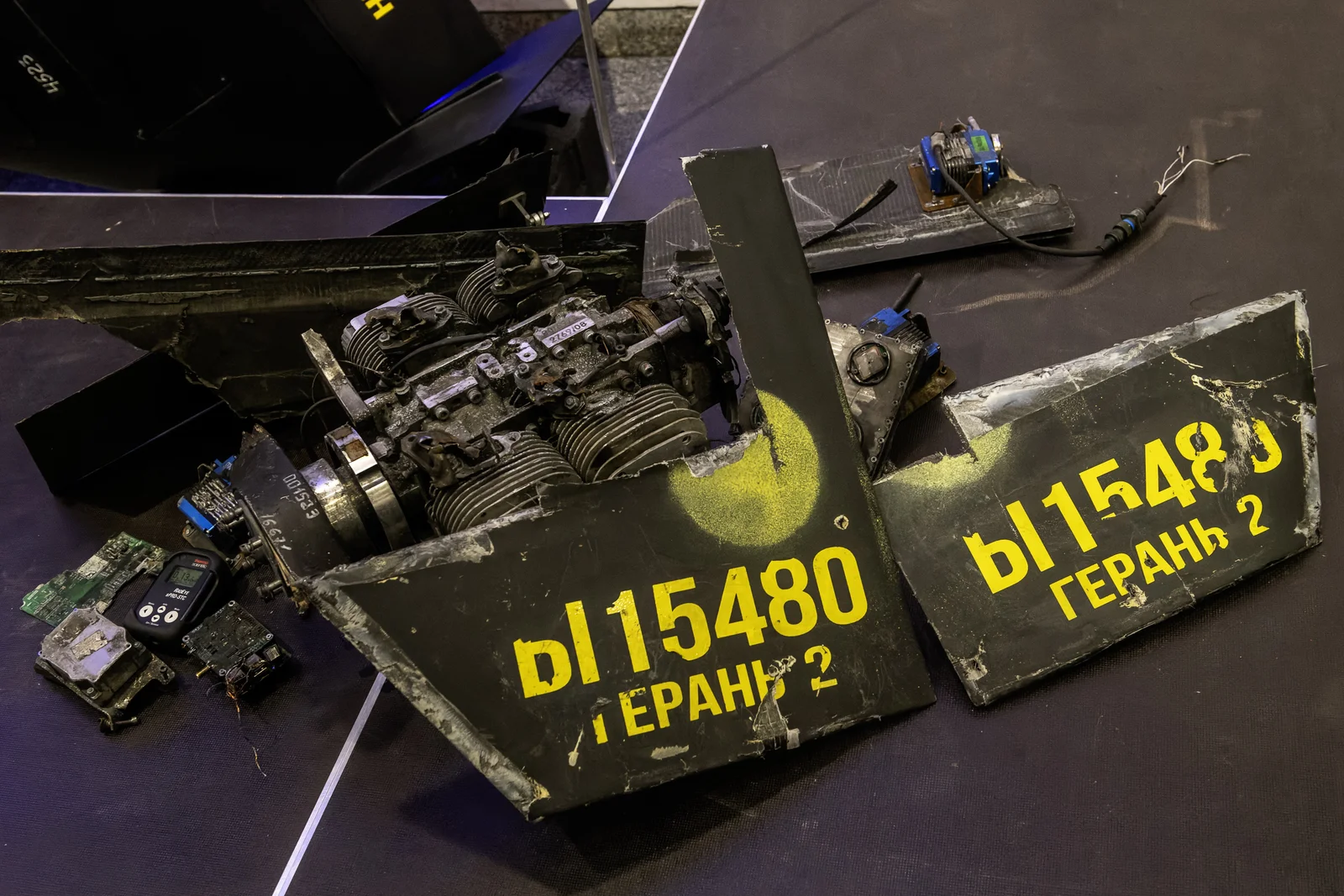 Recovered Geran-2 wreckage laid out on a display table, showing the four-cylinder two-stroke engine, control servos, circuit boards, and wing panels stenciled with the Cyrillic marking GERAN 2 and serial number 15480