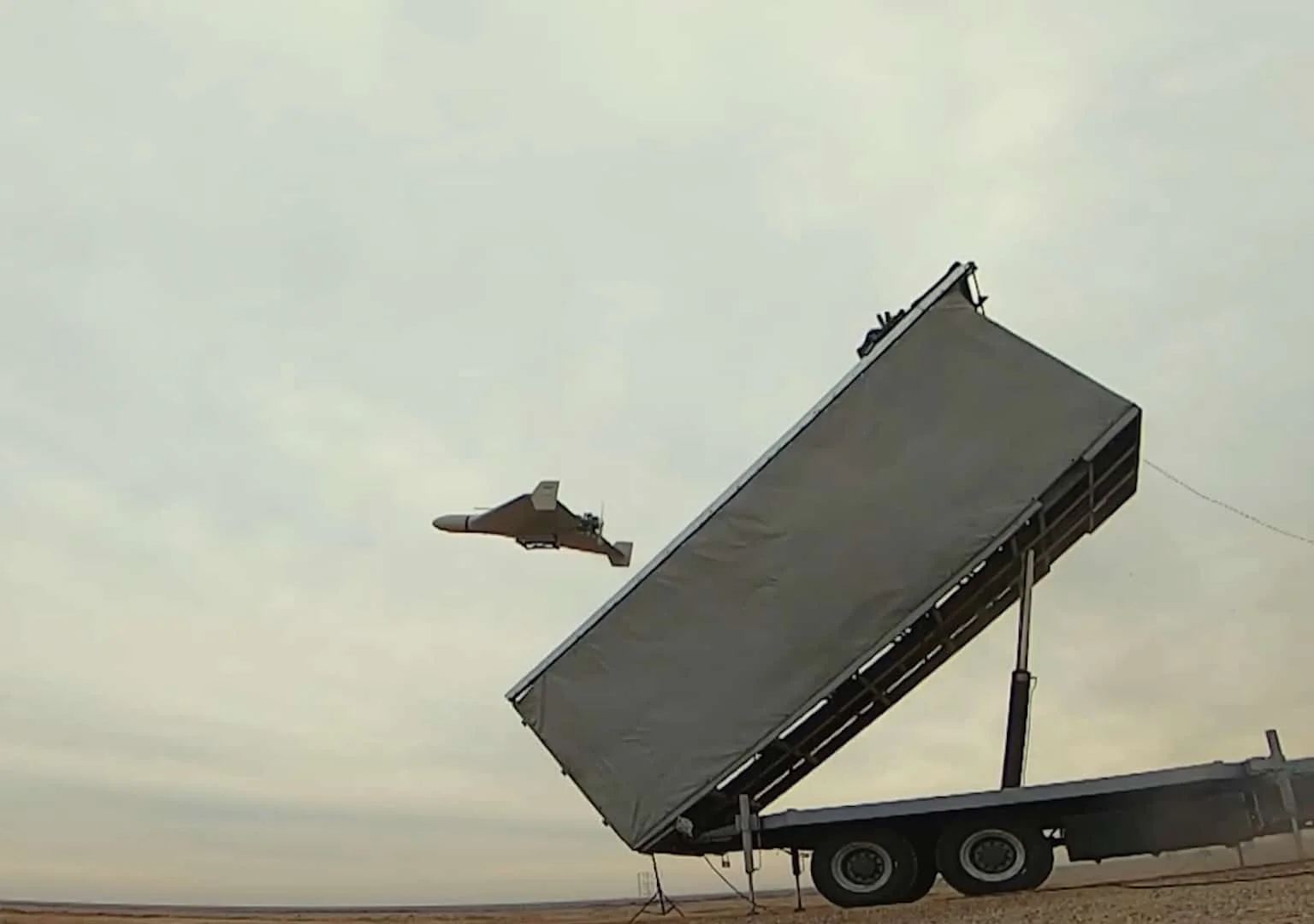 A Shahed-136 drone leaving a tilted truck-mounted launch rack under rocket boost against an overcast desert sky